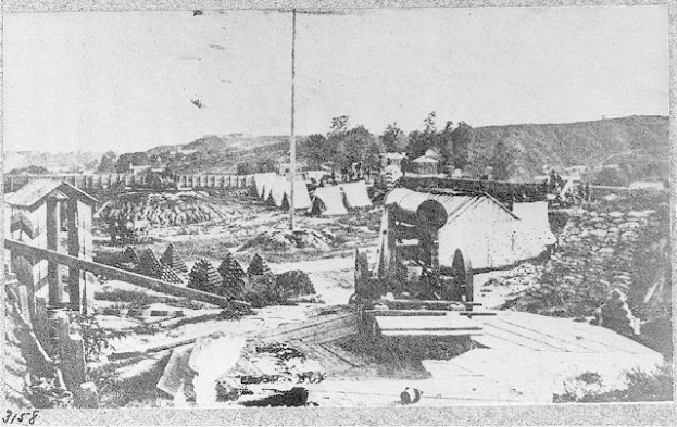 Schwarzes und weißes Foto einer Baustelle mit einer Maschine im Vordergrund, umgeben von Schuppen, Bäumen, Hügeln und einem Himmel im Hintergrund, mit Text unten, der lautet "Die Bauarbeiten an einer neuen Brücke in der Stadt San Francisco, Kalifornien."