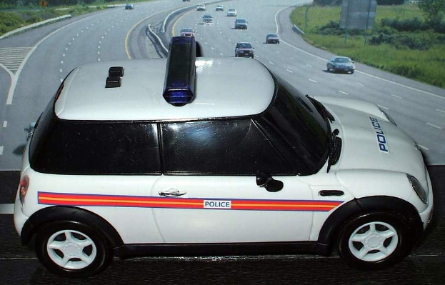 Weißer Polizeiwagen mit "Polizei" darauf vor einer Reihe von fahrenden Fahrzeugen auf einer von Gras und Bäumen gesäumten Straße außerhalb der Stadt.