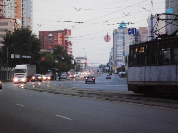Eine belebte Stadtstraße mit Verkehr, einschließlich einem Bus und Lastwagen, Straßenlaternen, Schildern, Bäumen, Strommasten mit Drähten, Gebäuden und einem klaren blauen Himmel.