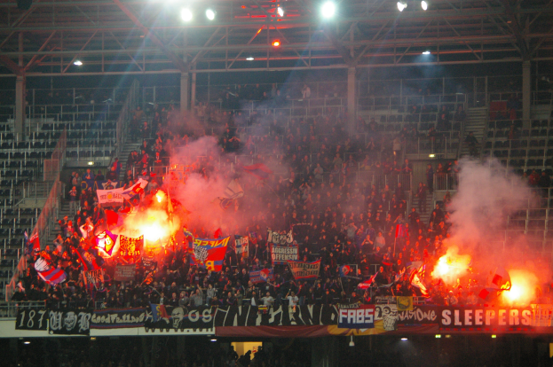 Eine große Menge Menschen in einem Stadion hält Fahnen und Banner, mit Leuchtraketen, die Rauch erzeugen, unter einer Decke mit Leuchten und Metallrahmen.