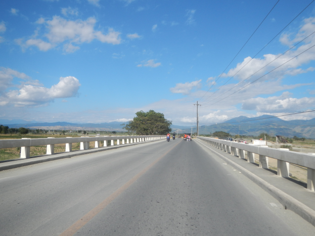 Eine Autobahn mit Motorradfahrern, umgeben von Geländern, Strommästen mit Käblein, Bäumen, Hügeln und einem bewölktem Himmel.
