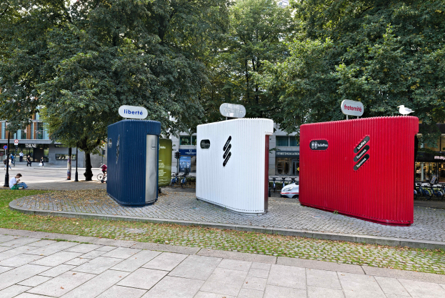 Drei mobile Toiletten an einer grasbewachsenen Straßenecke mit Gebäuden und einem klaren blauen Himmel im Hintergrund.