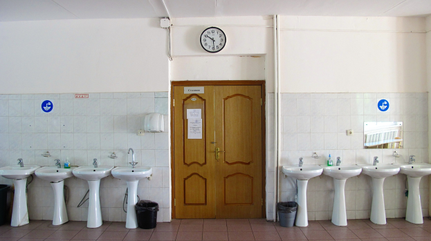 Reihe von Waschbecken mit Wasserhähnen, Bodenstaubsauger, eine Tür im Hintergrund, eine Wanduhr und ein Spiegel auf der rechten Seite in einem Badezimmer.