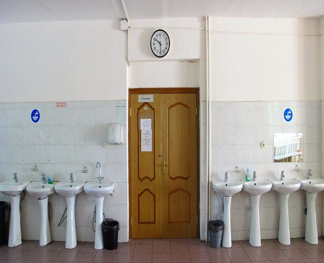 Reihe von Waschbecken mit Wasserhähnen, Bodenstaubsauger, eine Tür im Hintergrund, eine Wanduhr und ein Spiegel auf der rechten Seite in einem Badezimmer.