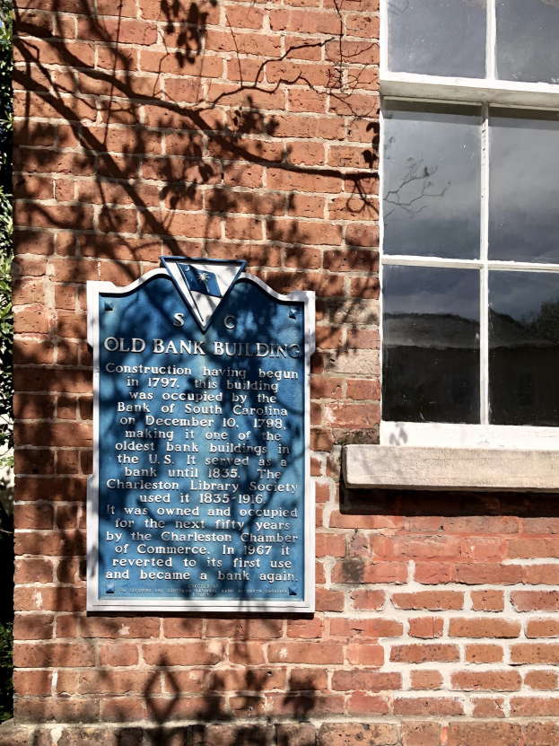 Altes Bankgebäude mit blauer Tafel an der Seite, Fenster auf der rechten Seite und Baum auf der linken Seite; die Tafel enthält Text.