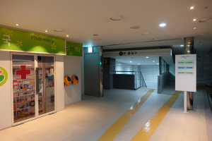 Apotheken-Eingang mit einem "Apotheke"-Schild auf der linken Seite, einer Tafel mit Text auf der rechten Seite, beleuchteten Lampen oben und undeutlichen Hintergrundobjekten.