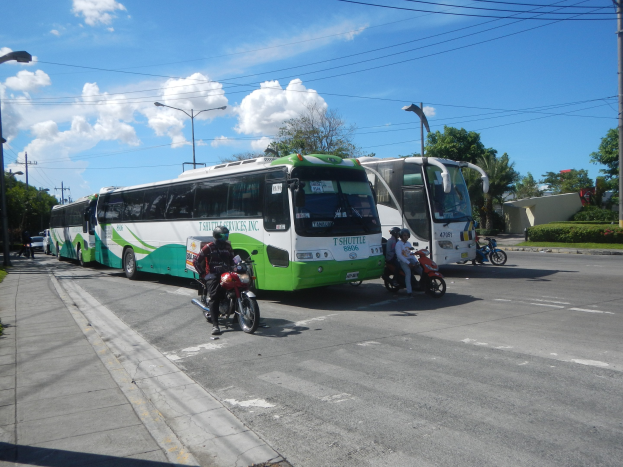 Ein grün-weißer Shuttlebus parkt am Straßenrand mit Motorrädern davor, einem grasbewachsenen Fußweg links und Gebäuden, Bäumen und Laternen im Hintergrund bei einem klaren blauen Himmel.