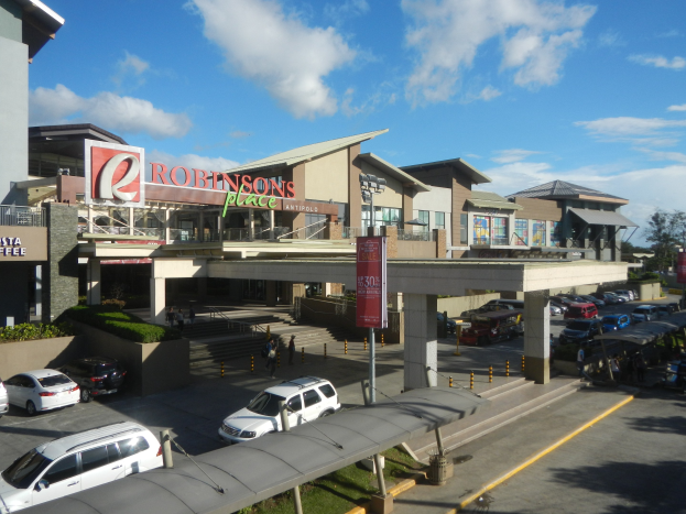 Ein belebter Einkaufszentrum mit Gebäuden, Schaufenstern, Fahrzeugen, Fußgängern, Straßenmöblierung, Grünflächen und einem bewölkten Himmel.