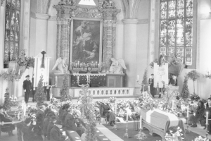 Schwarz-weißes Foto einer Trauerfeier in einer Kirche mit Sitzenden und Stehenden, Blumensträußen, Kerzen, einem Sarg im Vordergrund und einem Rahmenfoto und Skulpturen im Hintergrund.