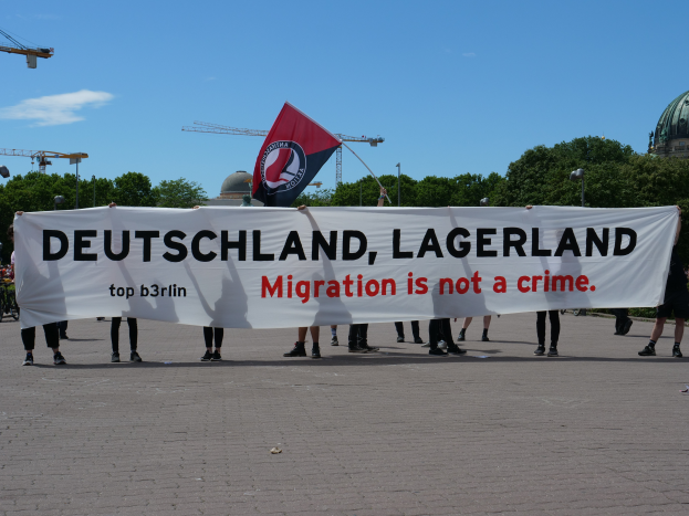 Eine Gruppe von Menschen mit einer Fahne, auf der "Deutschland, Lagerland Migration ist kein Verbrechen" steht, und einer Flagge, mit Straßenlaternen, Bäumen, Kränen, einem Gebäude und einem bewölkten Himmel im Hintergrund.