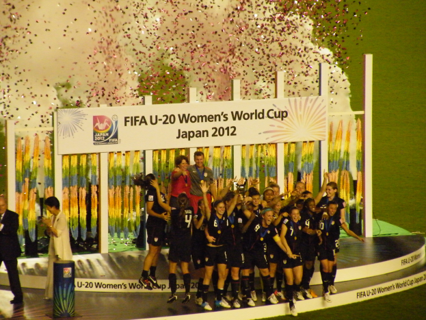 Eine Gruppe von Frauenfußballspielerinnen in schwarzen Uniformen steht auf einer Bühne, feiert ihren Sieg, umgeben von einer Menge mit Kameras, mit einem dekorierten Hintergrund und grasbewachsenem Boden.