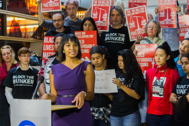 Eine Black-Lives-Matter-Aktivistin steht an einem Podium mit einem Mikrofon und spricht zu einer Menge, die Schilder hält, mit Scheinwerfern und Equipment im Hintergrund.