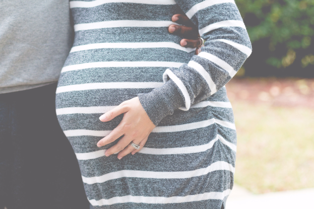 Schwangeres Paar steht nebeneinander mit den Händen auf dem Bauch, trägt Schwangerschaftskleidung und befindet sich in einer bewaldeten Outdoor-Umgebung.
