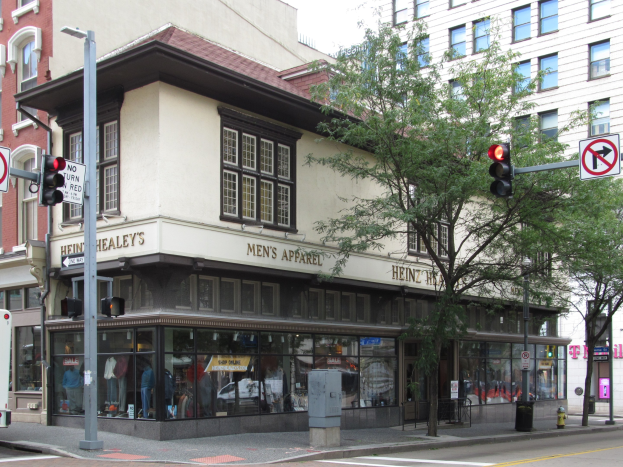Eine belebte Stadtstraße mit einem geparkten Bus, Gebäuden, Bäumen, Laternen, Verkehrsampeln, Schildern und einem Kaufhaus mit Schaufensterpuppen im Hintergrund unter einem sichtbaren Himmel.