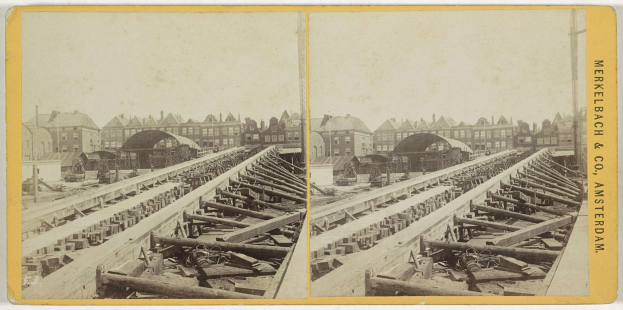 Schwarz-weiß-Foto einer Zugstrecke mit Gebäuden im Hintergrund, Text "Merkelbach & Co, Co. Amsterdam" auf der rechten Seite und verstreute hölzerne Gegenstände im Vordergrund.