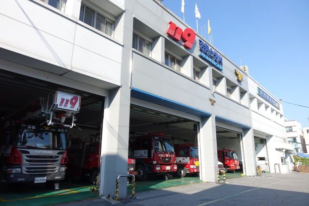 Feuerwache mit mehreren Feuerwehrautos vor dem Gebäude geparkt, mit Fenstern, Säulen und Namensschildern, sowie Fahnen im Vordergrund und anderen Gebäuden gegen einen klaren blauen Himmel.