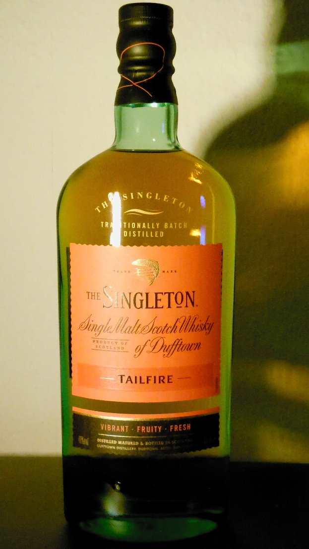 Eine tiefe bernsteinfarbene Flasche Singleton of Dufftown Tailfire Single Malt Scotch Whisky auf einem Tisch, mit einem schwarzen Etikett, das eine Löwen- und Burgkrone in Großbuchstaben und einem Wappen zeigt.