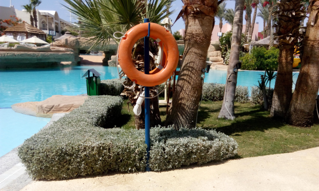 Ein Schwimmbad mit einem Rettungsring in der Mitte, umgeben von Grünflächen und Gebäuden unter einem klaren blauen Himmel.