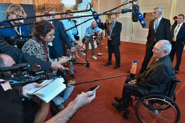 Ein Mann im Rollstuhl mit Anzug und Krawatte umgeben von Reportern mit Kameras und Mikrofonen, mit einem Banner und einer Tür im Hintergrund.