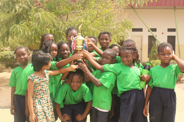 Gruppe von Kindern, die eine Trophäe in einem Schulhof mit Bäumen und einem Gebäude im Hintergrund halten.
