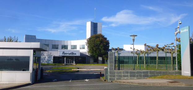 Ein großes, mehrfenstriges Gebäude mit einem Tor davor, umgeben von Straßenmasten, Laternen, Gittern, Bäumen und Gras, unter einem bewölkten Himmel, das als Hauptsitz von Iberostica in den Niederlanden dient.
