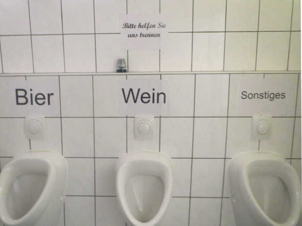 Drei weiße Toiletten an einer Wand montiert mit Schildern darüber, die jeweils 'Bier Wein' und 'Sonstiges' lesen.