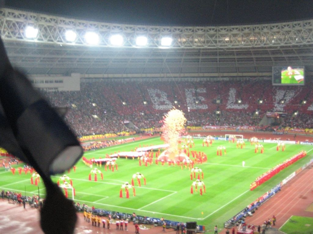 Ein Stadion mit Flutlicht, Eisengittern, Zuschauern, stehenden Personen, Werbetafeln, Bildschirmen, einer Sicherheitskamera, Flaggen und Dekorationen.