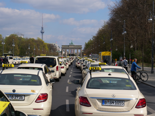 Eine lange Reihe von Taxis, die an der Seite einer belebten Straße in Berlin, Deutschland, geparkt sind, mit Fahrzeugen, Radfahrern und Fußgängern, flankiert von Laternenmasten, Bäumen und Gebäuden, einschließlich eines Bogens und eines Turms.