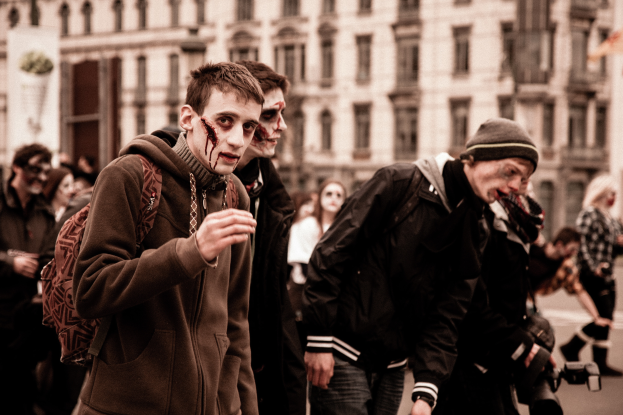 Eine Gruppe von Menschen in Zombie-Kostümen, die eine Straße entlanggehen, mit einem Gebäude im Hintergrund und einer Person mit einer Kamera.