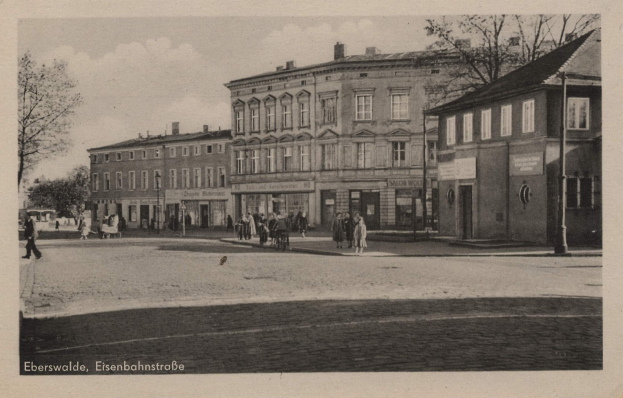 Schwarzes und weißes Foto von Eisenbahnstraße in Eberswalde mit Gebäuden, Bäumen, Fußgängern, Strommasten und Schildern unter einem bewölkten Himmel, mit Text unten.