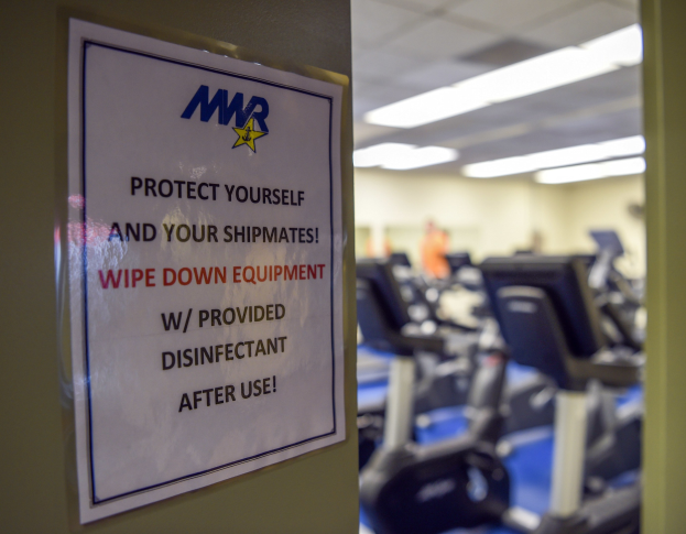 Ein Türschild mit der Aufschrift "Schützen Sie sich und Ihre Schiffskameraden" mit Laufbändern und Deckenleuchten im Hintergrund, Hintergrund leicht unscharf.