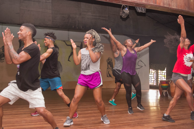 Gruppe von Menschen, die in einem Studio mit einer bemalten Wand, einem Fenster und Deckenbeleuchtung tanzen, was auf eine Zumba-Stunde hinweist.