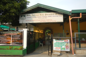 Der Eingang der San Lorenzo Ruiz Pfarrschule zeigt ein Schild mit Text, einen Metallzaun, Menschen auf Stühlen unter einem Dach, Pfosten, ein Schild, Pflanzen, einen Baum und einen bewölkten Himmel.