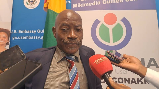 Ein Mann im Anzug spricht mit Reportern, während im Vordergrund eine Person mit einem Mikrofon und einem Gerät zu sehen ist. Im Hintergrund befinden sich Schilder, Fahnen und eine Frau.