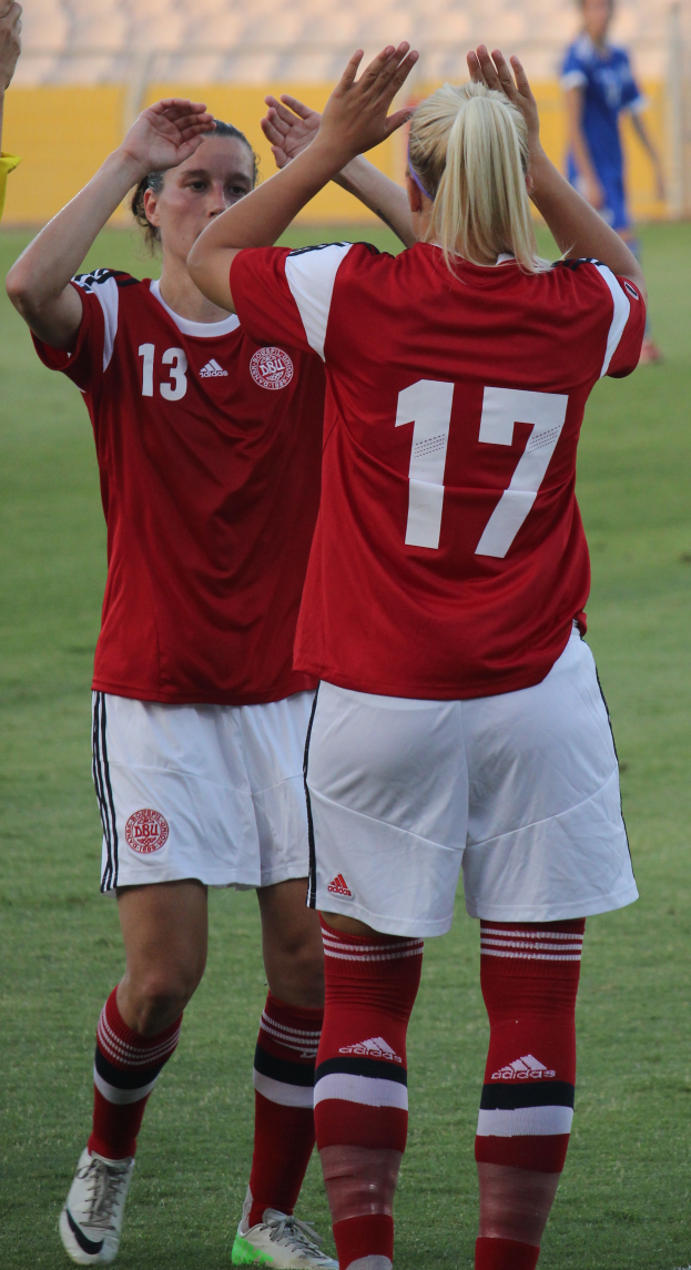 Zwei Frauen in roten T-Shirts und weißen Shorts stehen auf einem Fußballfeld und klatschen in die Hände, während im Hintergrund eine Person in einem blauen Kleid, eine gelbe Tafel und einige Stühle zu sehen sind.