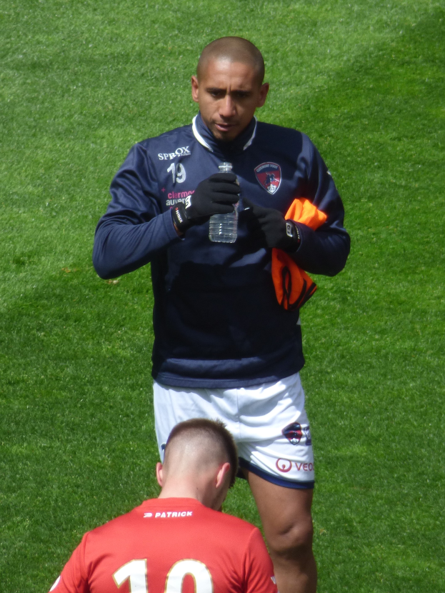 Ein Mann in einem Sportoutfit steht auf einem grünen Feld und hält eine Flasche in einer Hand und ein oranges Tuch in der anderen.
