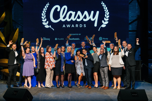 Eine Gruppe von Menschen auf einer Bühne, die Auszeichnungen halten, mit Rednern darunter und einem Bildschirm im Hintergrund, der "Classy Awards 2019" anzeigt.