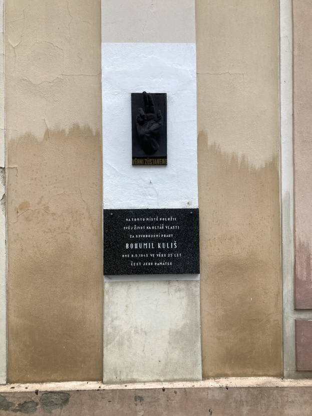Schwarze Tafel mit eingravierter Schrift an einer Gebäudewand, eine Hand ruht darauf, wahrscheinlich ein Holocaust-Mahnmal.