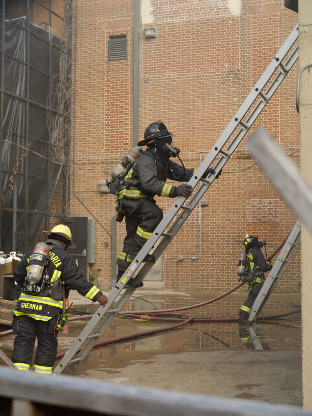 Feuerwehrleute in Helmen und Lufttanks, die eine Leiter vor einem Backsteingebäude mit Rohren auf dem Boden und einer Metallstange am Boden hinaufklettern, mit einem weiteren Gebäude mit Fenstern und einem Netz im Hintergrund.