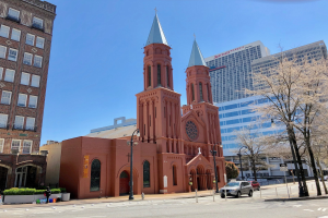 Die St. Patrick's Cathedral, eine große rote Kirche mit einem Turm, steht in der Mitte einer Straße in Philadelphia, Pennsylvania, umgeben von Gebäuden, Straßenlaternen, Straßenlaternen, Kraftfahrzeugen, Fußgängern, Büschen, Bäumen und unter einem klaren blauen Himmel.