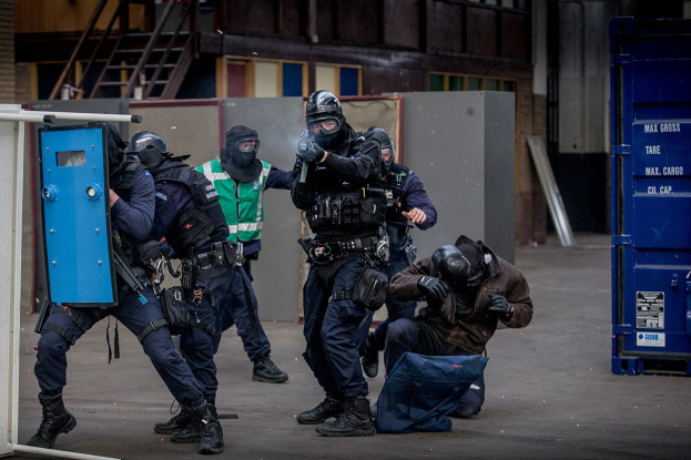 Polizeibeamte in Einsatzausrüstung stehen vor einem Gebäude, mit einer Person am Boden im Vordergrund und einem blauen Objekt mit Text auf der rechten Seite.