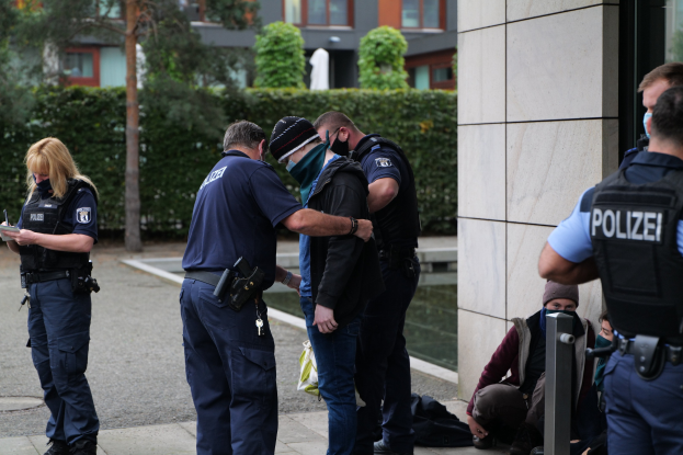 Eine Gruppe von Polizisten steht vor einem Gebäude, während eine Frau ein Buch und einen Stift hält, mit sitzenden und stehenden Personen im Hintergrund bei Bäumen und Gebäuden.