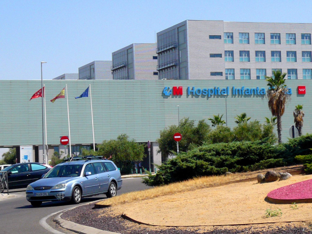 Krankenhaus mit parkenden Autos, umgeben von urbaner Infrastruktur und Vegetation unter einem klaren blauen Himmel.