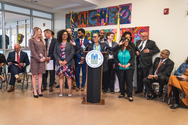 Eine Gruppe von Menschen steht vor einem Rednerpult mit einem Mikrofon und einer Tafel mit der Aufschrift "Sancho City Councilman Joins City Council", mit anderen im Hintergrund sitzend und stehend, Flaggen, eine Tafel, ein Feuerlöscher und Deckenleuchten an der Rückwand.