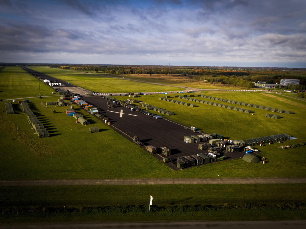 Luftaufnahme einer Militärbasis in den Niederlanden mit Fahrzeugen auf der Straße, Grasflächen, Bäumen, Gebäuden und einem bewölkten Himmel.