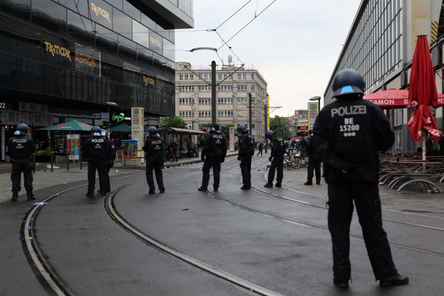 Polizeibeamte in schwarzen Uniformen und Helmen stehen entlang einer Straße, die von Glasfenstergebäuden gesäumt ist, mit Bäumen, Strommasten und verstreuten Außenmöbeln unter einem klaren Himmel.
