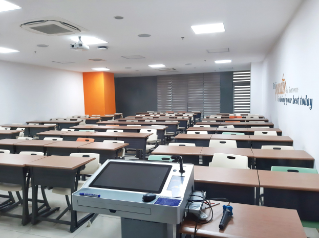 Ein Klassenzimmer mit Tischen und Stühlen in Reihen, einem deckengehängten Projektor und Gegenständen auf den Tischen, mit Text an der rechten Wand, der darauf hinweist, dass es eine Privatschule in Singapur ist.