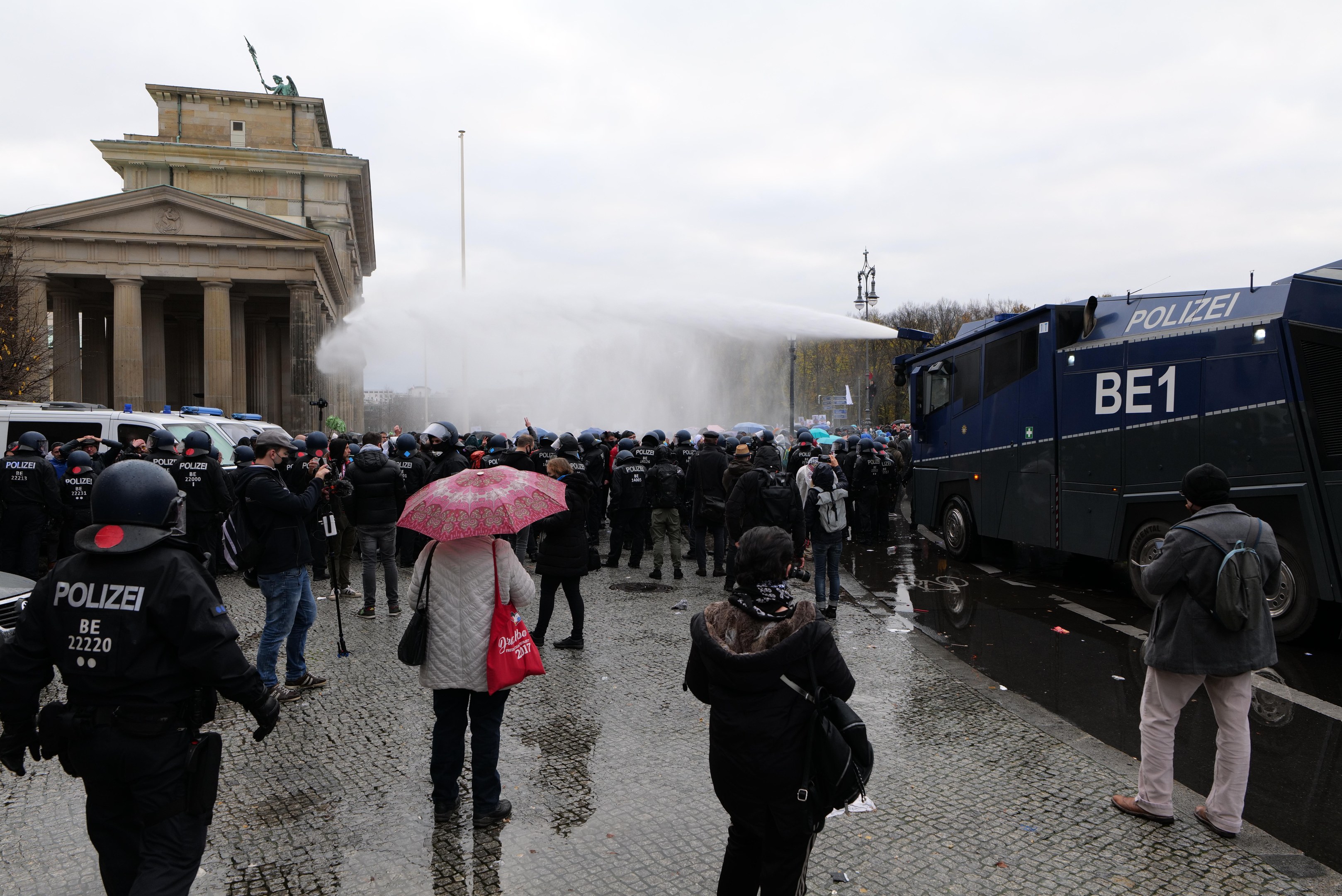 Eine Gruppe von Menschen, die vor einer Reihe von Polizeibeamten in Berlin steht, einige tragen Helme und halten Schirme, mit Fahrzeugen, einem Gebäude mit Säulen, Bäumen und Laternen im Hintergrund.