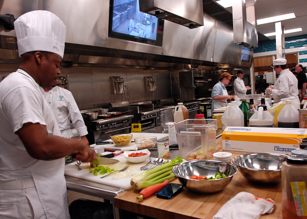 Eine Gruppe von Köchen in einer Küche, die Essen auf einem Schneidbrett zubereiten, mit einem Koch im Vordergrund in einer weißen Schürze und Mütze, der ein Messer hält, umgeben von verschiedenen Utensilien, Schüsseln, Gemüse, einem Handy, Flaschen und anderen Gegenständen, mit einem Fernseher und Deckenleuchten im Hintergrund.