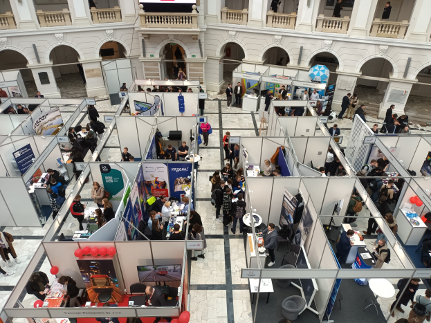 Eine große Gruppe von Menschen auf einer internationalen Fachmesse mit Ständen, Bannern, Tischen, Stühlen und Luftballons in einem Raum mit architektonischen Bögen und Säulen.